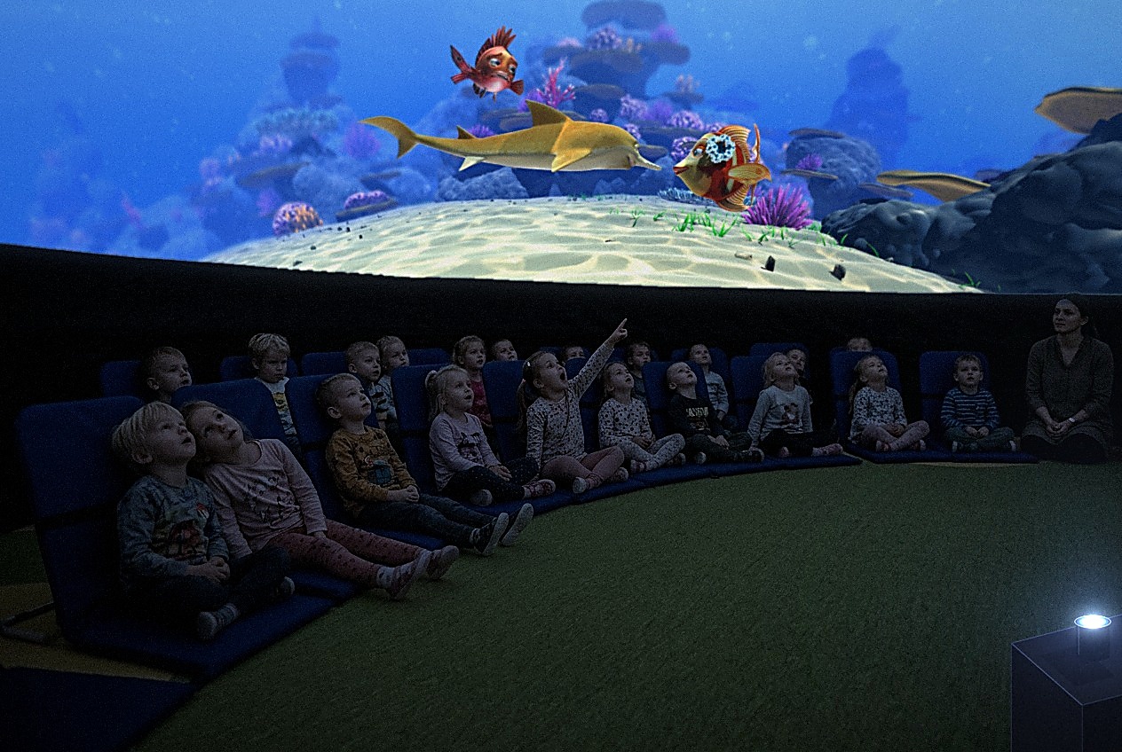 Dzieci z przedszkola siedzą na krzesełkach z oparciem w kopule planetarium i oglądają film o podwodnym świecie